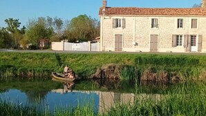 Un bâtiment en pierre avec un toit en tuiles, une zone herbeuse et une étendue d’eau calme dans laquelle se reflète la structure.