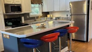 Une cuisine moderne avec des armoires blanches, un réfrigérateur en acier inoxydable, un four à micro-ondes et une cuisinière à gaz. On y trouve trois tabourets de bar avec des assises bleues et rouges.
