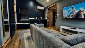 A modern living room with a sectional sofa, a flat-screen TV, and a kitchen area with a window and blinds. - Lone Ranger RV Park & Lodge (Ranger)