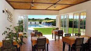 Un comedor interior con mesas y sillas, una piscina visible a través de puertas de cristal y vistas al exterior.