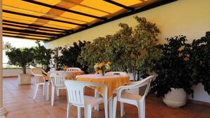 Un patio al aire libre con sillas y mesas de plástico blanco, rodeado de plantas en macetas y un suelo de baldosas.