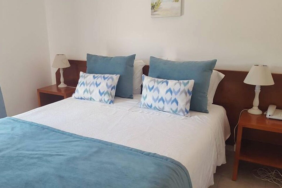 A neatly made bed with a blue blanket and patterned pillows, two bedside lamps, and a small bedside table with a phone.