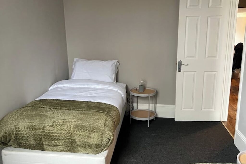 A small, neatly arranged bedroom with a single bed, a bedside table, and a door leading to another room.