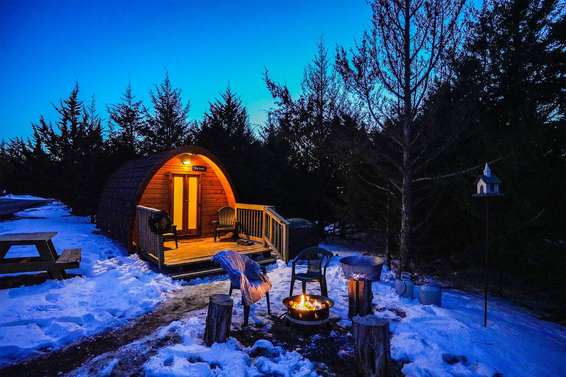 Una cabaña acogedora de troncos, con luz cálida en el interior, un brasero y un banco en el exterior.