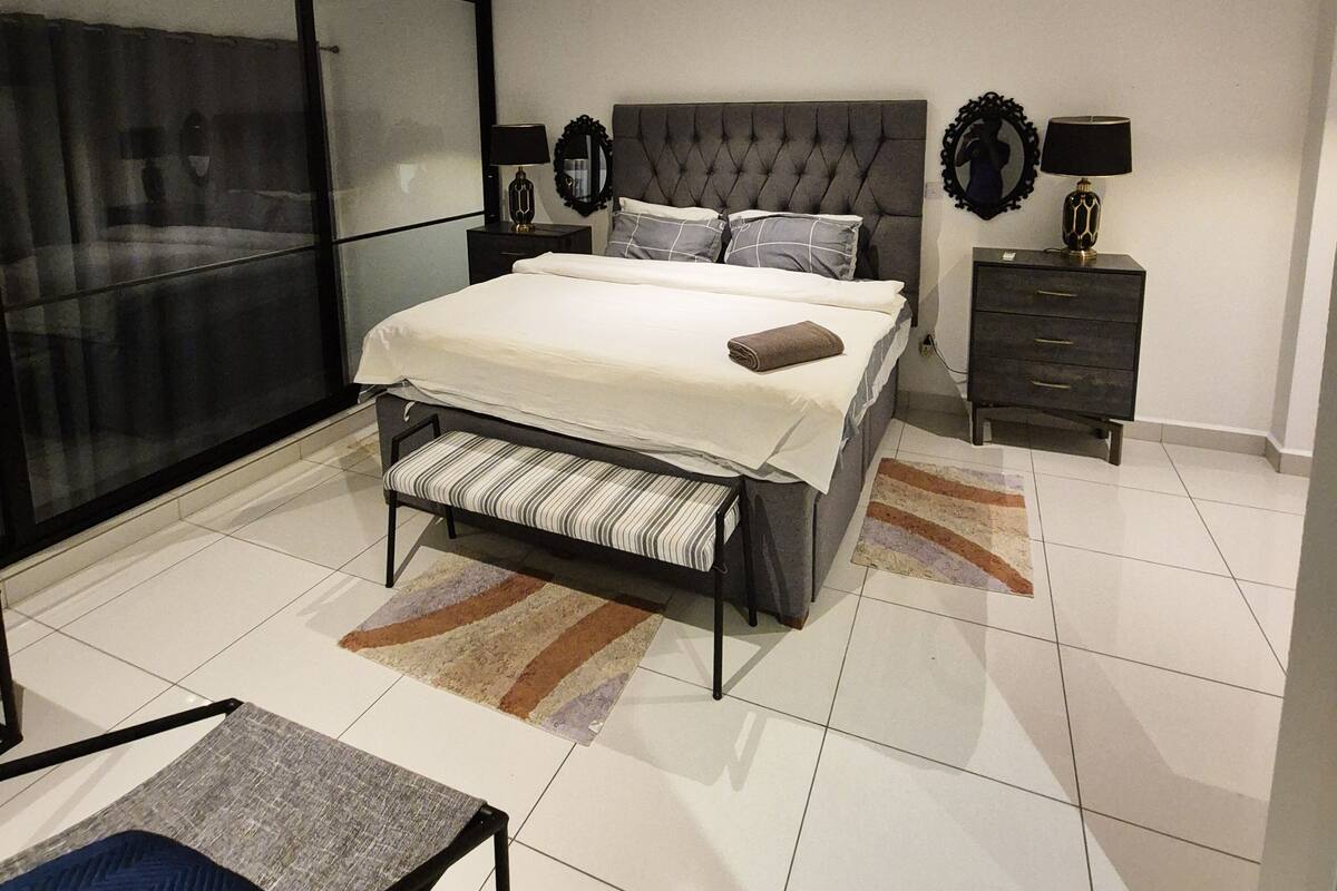 A modern bedroom with a large bed, a bench, a nightstand, and a mirror.