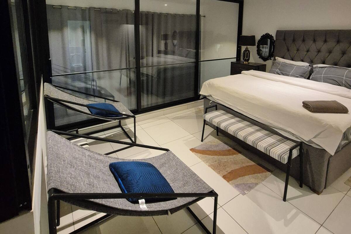 A modern bedroom with a large bed, a glass partition, a sofa, and a bench.