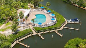 Eine Vogelperspektive zeigt ein Resort mit einem runden Pool, umgeben von Liegestühlen und Sonnenschirmen, verbunden mit dem Fluss durch eine Brücke.