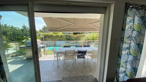 Une porte-fenêtre coulissante donne sur une terrasse extérieure équipée d’une piscine, d’un barbecue et d’une table avec des chaises.