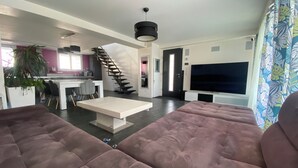 Un salon moderne doté d’un grand canapé d’angle, d’un coin repas avec une table et des chaises blanches, d’une cuisine avec un mur d’accent violet et d’un escalier menant à un autre niveau.