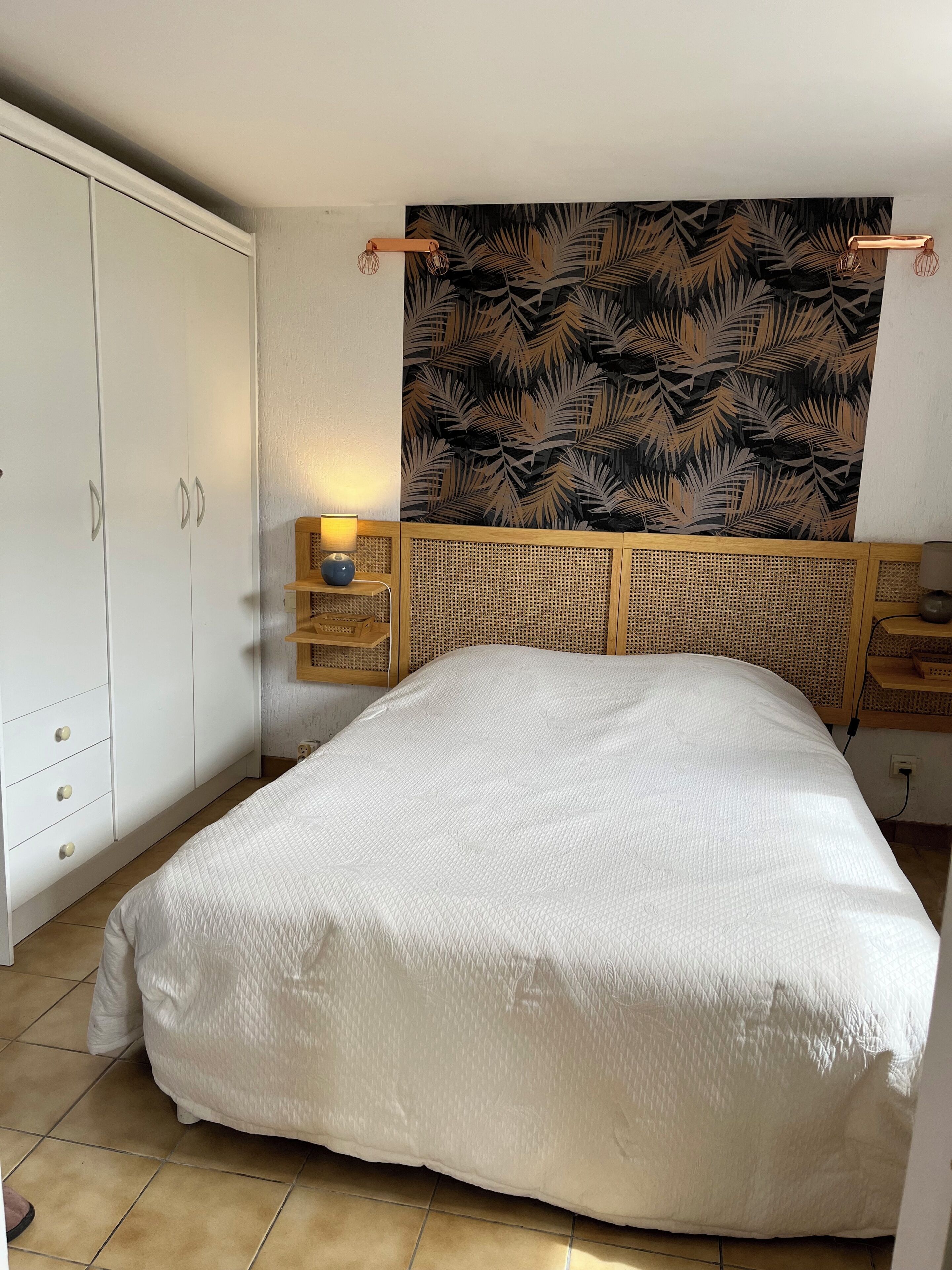 A bedroom with a bed, a wardrobe, a bedside table with a lamp, and a wall with a leaf pattern.