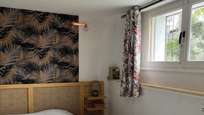 Une chambre à coucher avec un lit, une tête de lit en bois, une table de chevet avec une lampe, une fenêtre avec des rideaux et un mur recouvert d’un papier peint à motifs de feuilles.