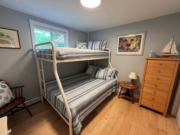 A bunk bed with striped bedding, a wooden dresser, a small table with a lamp, and a framed painting on the wall.