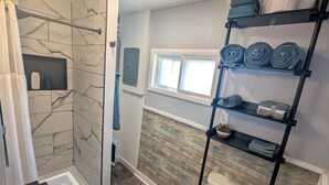 A bathroom with a shower, toilet, and sink. There is a window, a shelf with towels, and a shelf with toiletries. - Farmhouse Studio just minutes from uptown. (Butte)