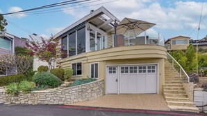 Une maison moderne à deux étages, dotée d’un mur de soutènement en pierre, d’un garage et d’un escalier menant à un balcon.