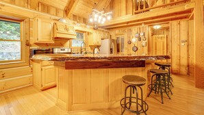 A wooden cabin interior with a kitchen featuring wooden cabinets, a large island with stools, and a view of the outdoors through a window. - Log Cabin w/ Vaulted Great Room & Creek Near GSMNP (Sevierville)
