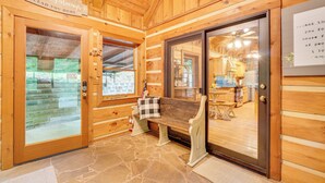 A wooden cabin interior with a stone floor, a bench, and a glass door leading to a kitchen area. - Log Cabin w/ Vaulted Great Room & Creek Near GSMNP (Sevierville)