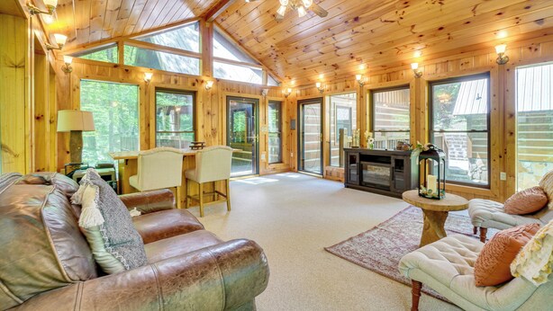 A spacious cabin interior with wooden walls, large windows, a fireplace, leather sofas, and a dining area. - Log Cabin w/ Vaulted Great Room & Creek Near GSMNP (Sevierville)