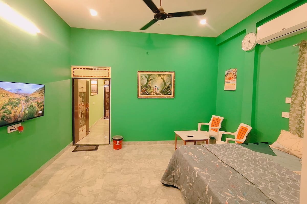 A room with green walls, a bed, a ceiling fan, a TV, and a door leading to another room.