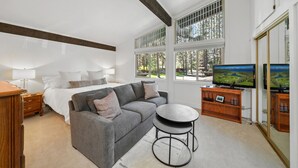 Ein Schlafzimmer mit Bett, grauem Sofa, rundem Couchtisch, Nachttisch aus Holz und einem Fernseher auf einem Holzständer.