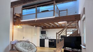 A loft-style living space with a high ceiling, exposed wooden beams, a dining area with a round table and chair, a kitchen with modern appliances, and a large flat-screen TV. - Domaine Villadomar: Farmhouse nestled in the heart of Thuir in the calm of its wooded grounds (thuir)