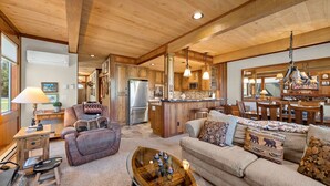 A spacious living room with a large sofa, a coffee table, and a fireplace. There is a kitchen area with wooden cabinets and a refrigerator.