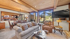A cozy living room with a sofa, armchair, and coffee table, a dining area with chairs and a table, and large windows offering a view of the outdoors.