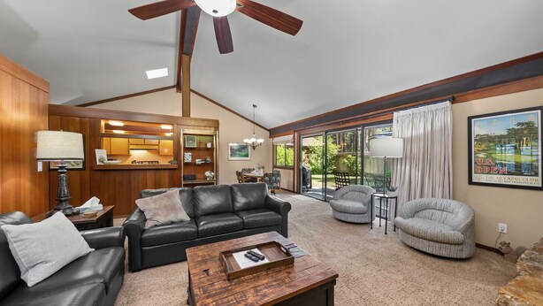 A spacious living room with a black leather sofa, a wooden coffee table, and a ceiling fan. There is a dining area with a kitchen in the background, and a large window with curtains. - Nestled in the heart of Graeagle Meadows, Private Fenced Yard, Sleeps 6 (Graeagle)
