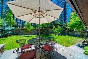A patio with a round table and chairs, a large umbrella, and a view of a well-maintained lawn and trees. - Sophisticated Remodel, Single Level, Fenced Yard (Graeagle)