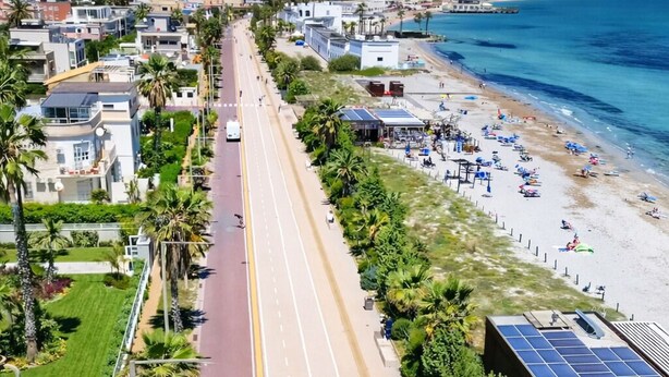 Une route côtière, avec une plage d’un côté, bordée de palmiers et de bâtiments.