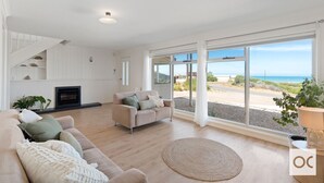 A spacious living room with a beige sofa, a round rug, and a fireplace. Large windows offer a view of the beach and ocean.