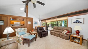 A living room with a brown leather sofa, a floral armchair, a wicker ottoman, a wooden coffee table, and a ceiling fan.