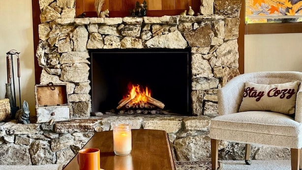 A stone fireplace with a fire, a wooden table with a candle, and a chair with a pillow. - Vacation retreat for golf and enthusiasts and families alike! (Graeagle)