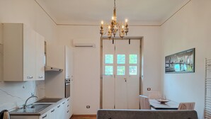 Eine moderne Küche mit weißen Schränken, einem eingebauten Backofen und einer Edelstahl-Spüle. Es gibt einen Essbereich mit Tisch und Stühlen sowie eine Sitzecke mit einem Kissen mit Leopardenmuster. Eine Kristallleuchter hängt von der Decke, und ein Gemälde ist an der Wand.