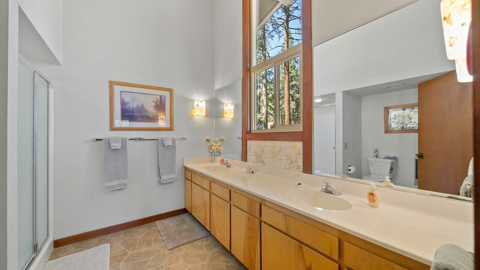 A bathroom with a large mirror, two sinks, wooden cabinets, and a window with a view of trees.