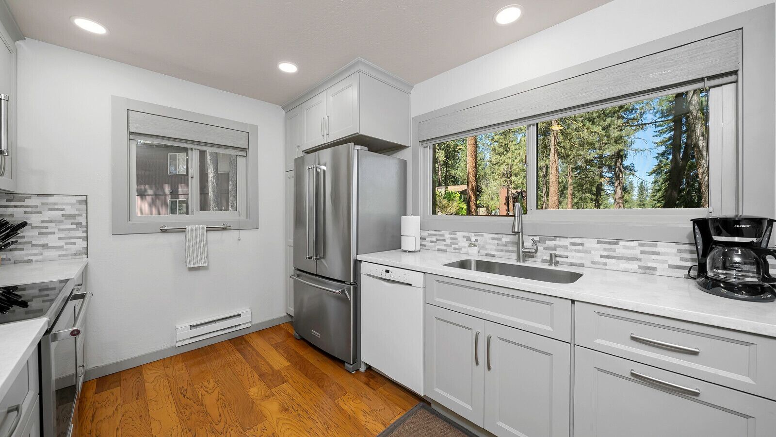 A modern kitchen with white cabinets, stainless steel appliances, a window with a view of trees, and a coffee maker on the counter.