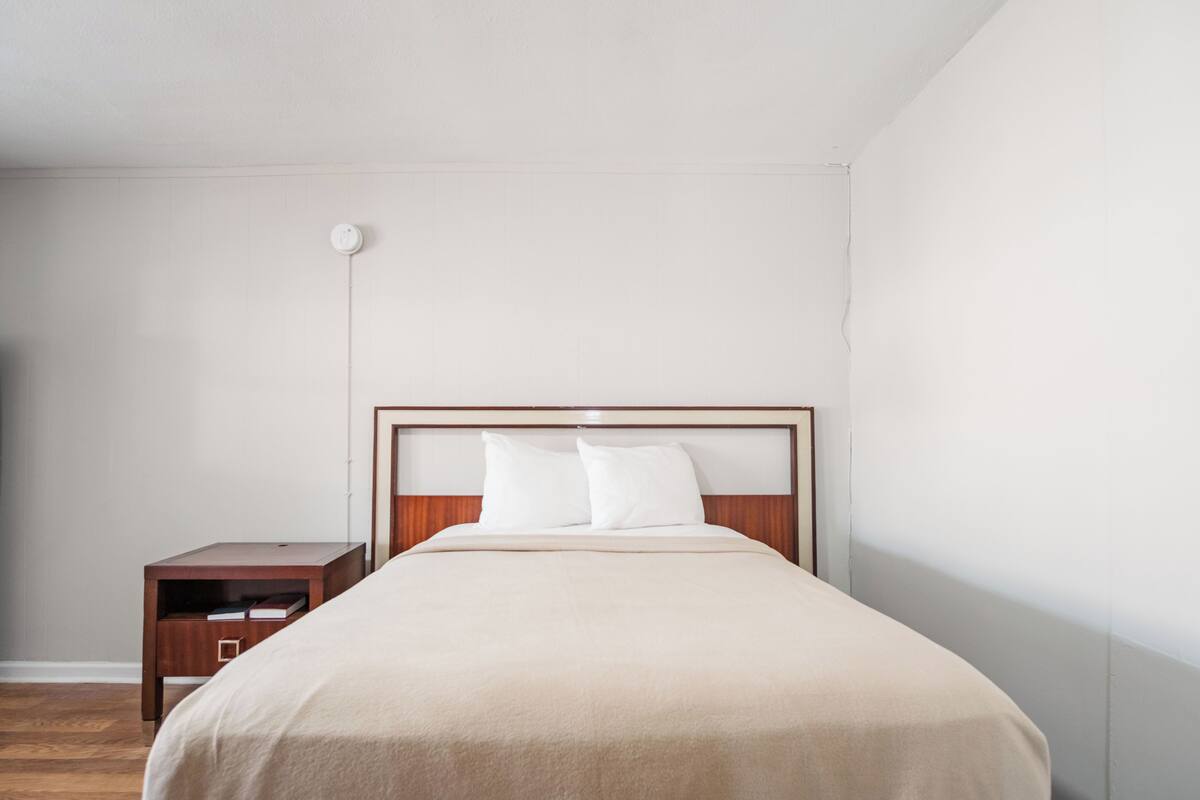 Una cama bien hecha con un cabecero de madera en una habitación sencilla, con una mesita de noche de madera cerca.