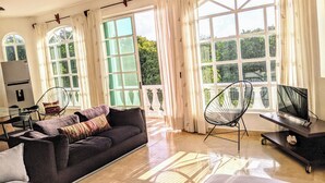 Amplia sala de estar con un sofá oscuro, un televisor sobre un mueble bajo y grandes ventanas con cortinas.