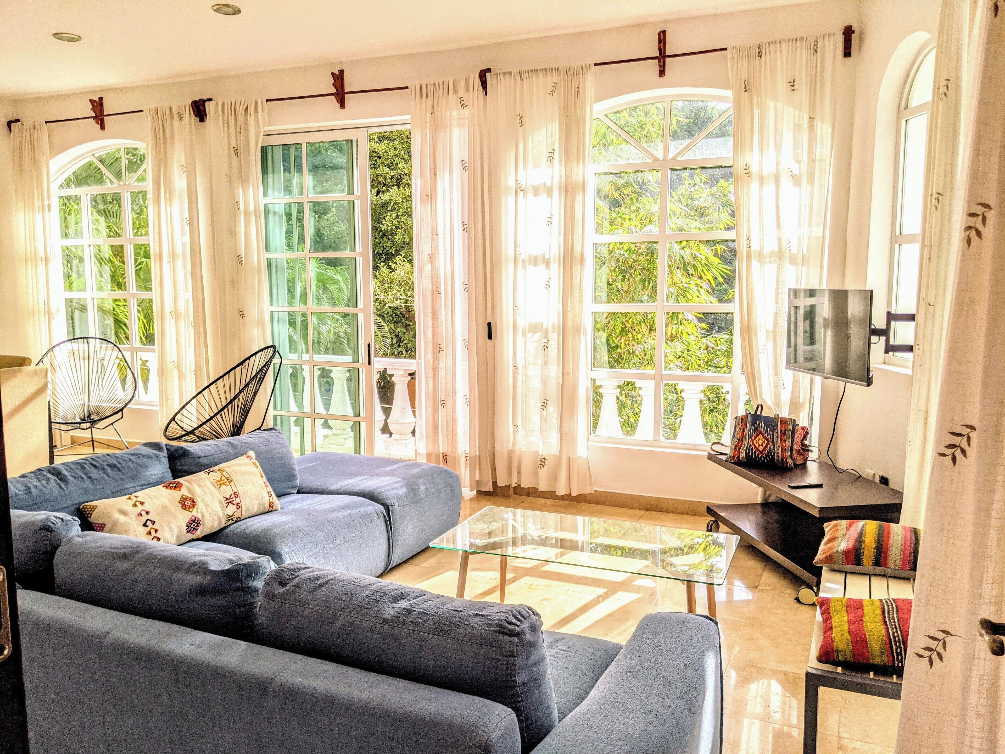 Un salon avec un canapé bleu, une table basse en verre et un téléviseur posé sur un meuble. De grandes fenêtres sont ornées de rideaux, et on y trouve un fauteuil ainsi qu’une petite table sur laquelle reposent un sac et une télécommande.