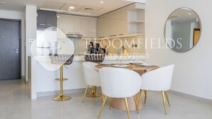 A modern kitchen with white cabinets, a round dining table with white chairs, and a gold-colored bar stool. - Bloomfields Grandeur 1BR In Lamar Raha (Abu Dhabi)