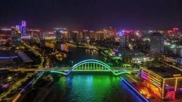 Paisaje urbano nocturno con un puente muy iluminado sobre un río, rodeado de edificios iluminados y un área costera bien iluminada.