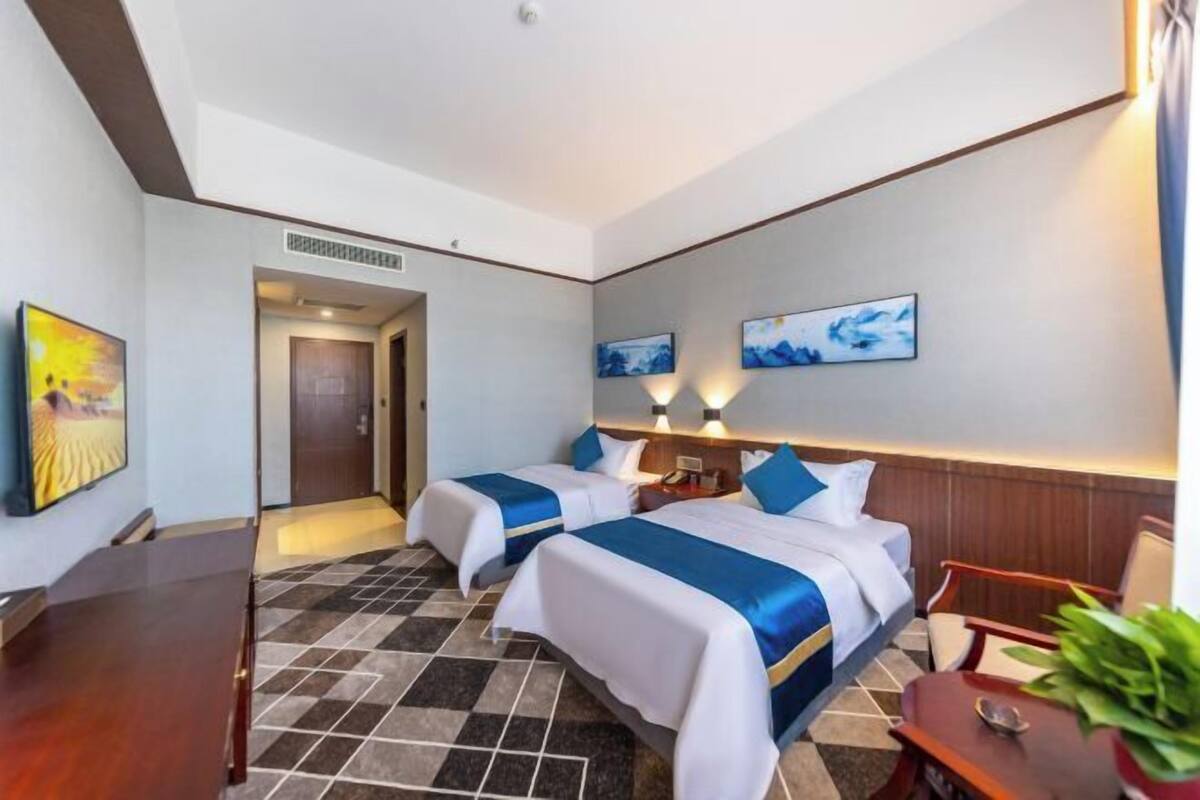 A hotel room with two beds, a desk, and a chair. There are two paintings on the wall and a plant on the desk.