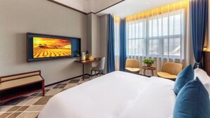 Ein Hotelzimmer mit einem Bett, einem Schreibtisch, zwei Stühlen, einem an der Wand befestigten Fernseher und einem Fenster mit Vorhängen.