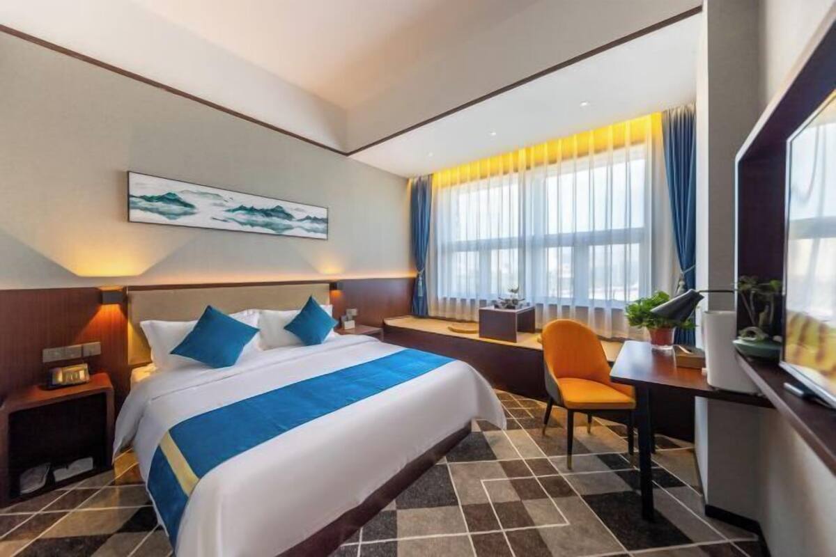 A hotel room with a large bed, a desk, a chair, a window with curtains, a painting on the wall, and a nightstand with a lamp.