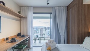 Hotellihuone, jossa on sänky, työpöytä tuolilla, pieni pöytä ja suuret ikkunat, joista avautuu näkymä kaupunkiin.