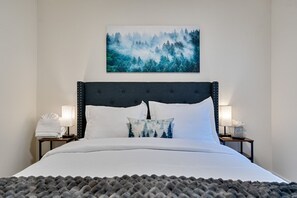 A neatly made bed with white linens, a dark headboard, and a forest-themed pillow. There are two bedside lamps on either side of the bed.