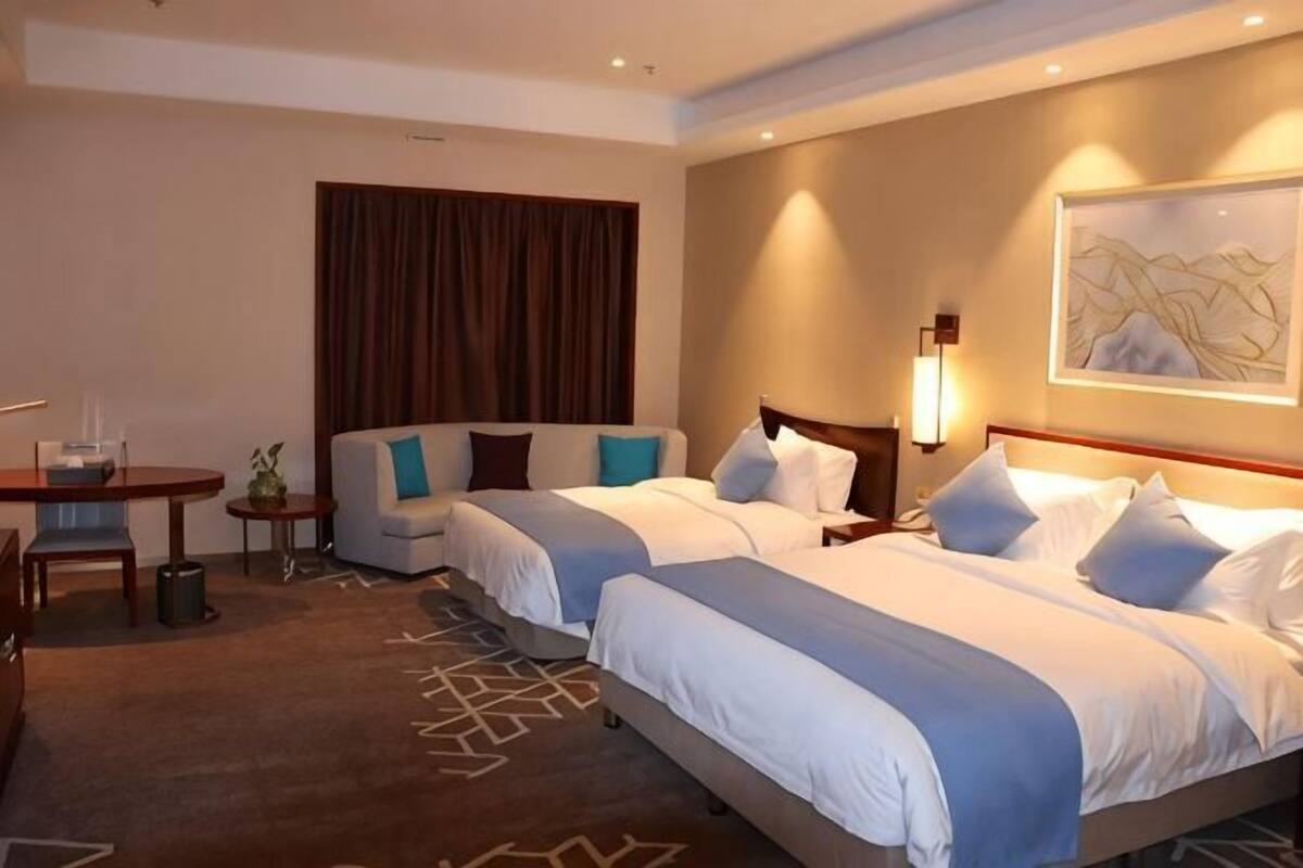 Une chambre d’hôtel avec deux lits, un bureau, une chaise, une petite table, une lampe et un tableau au mur.