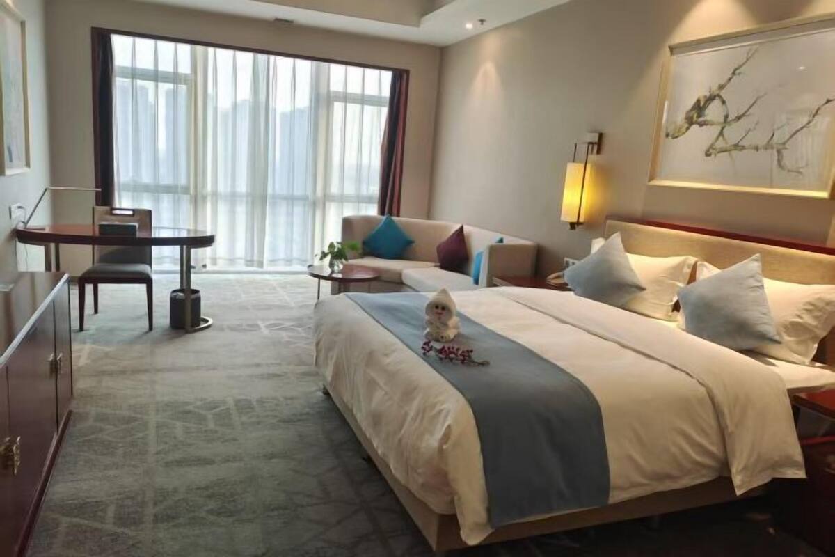 Une chambre d’hôtel avec un grand lit, un bureau, une chaise, un canapé et une grande fenêtre.