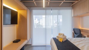 Moderni hotellihuone, jossa on sänky, hyllyllä oleva televisio, ikkunasta avautuva näkymä kaupunkiin sekä työpöytä ja tuoli.