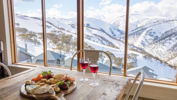 Una mesa con una tabla de quesos y dos copas de vino tinto, frente a una vista nevada de montañas a través de amplios ventanales.