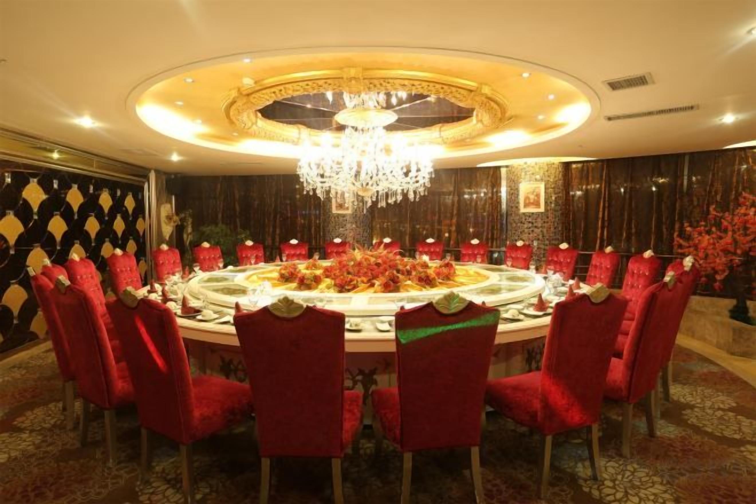 A round dining table with red upholstered chairs, surrounded by a chandelier and a patterned wall.